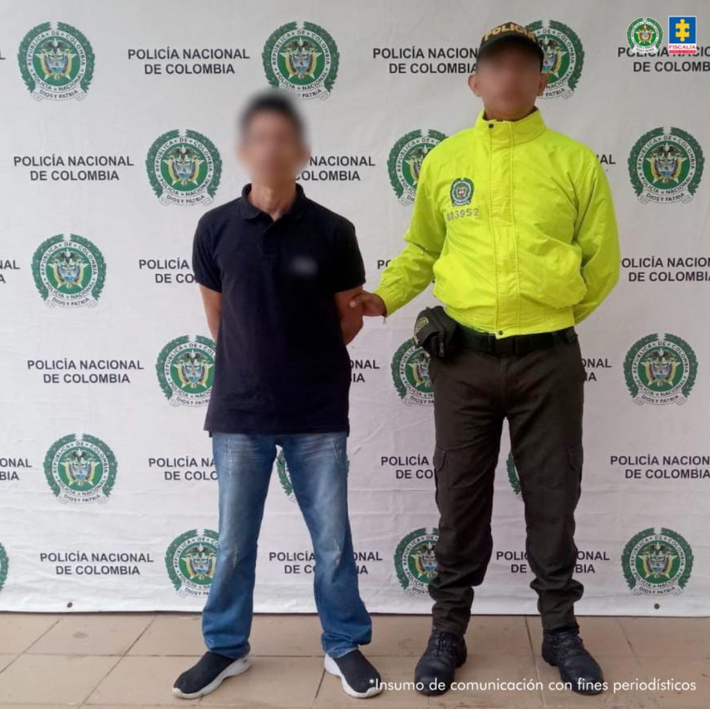 En la fotografía aparece un hombre de pie, con el rostro difuso, los brazos atrás, de estatura media, vestido con camiseta azul oscura, jean azul y tenis negros, custodiado por un uniformado de la Policía Nacional.